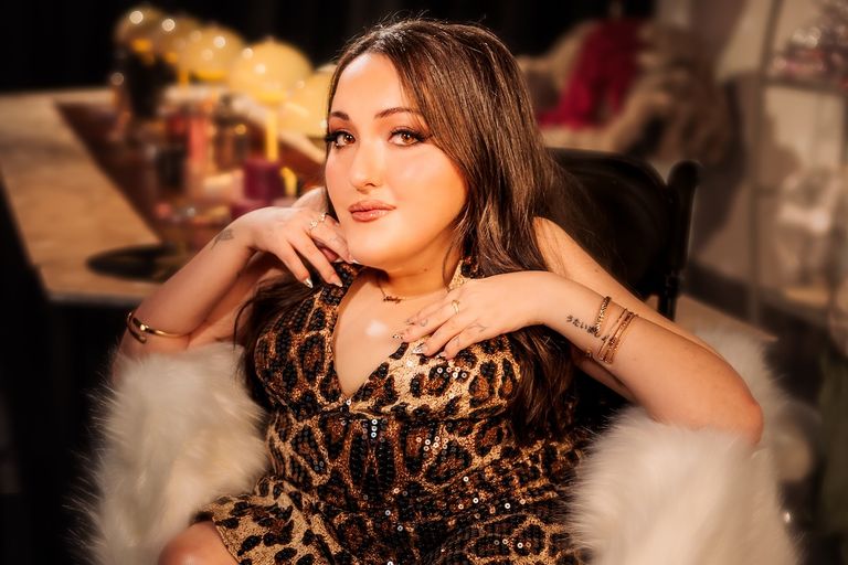 Glamorous promo portrait of artist Nat Luna seated in a leopard-print sequined dress with warm ambient lighting.
